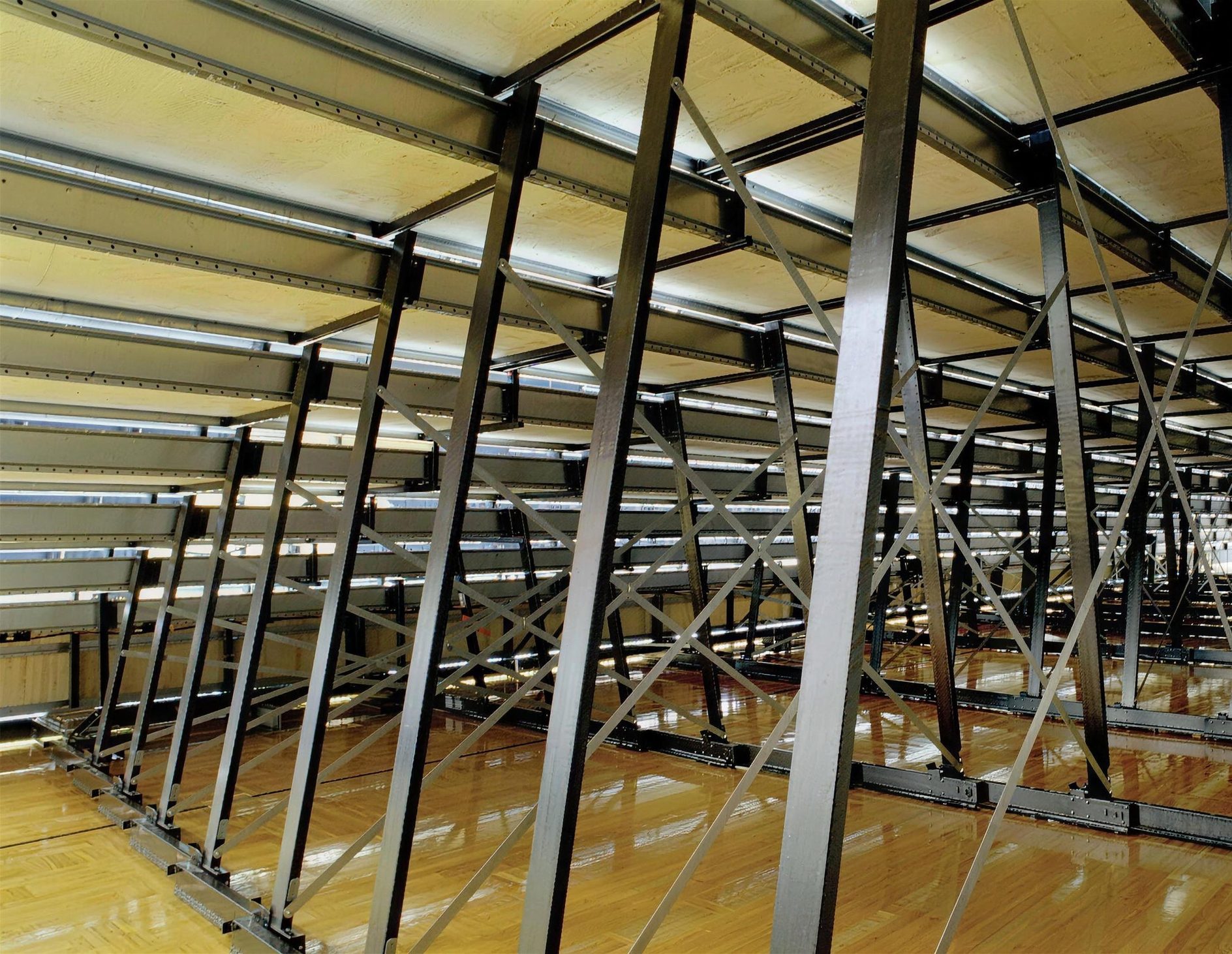 Underside of a MAXAM telescopic gym seating bank, showing slant frames, tension bracing and full-length deck support over a hardwood floor.